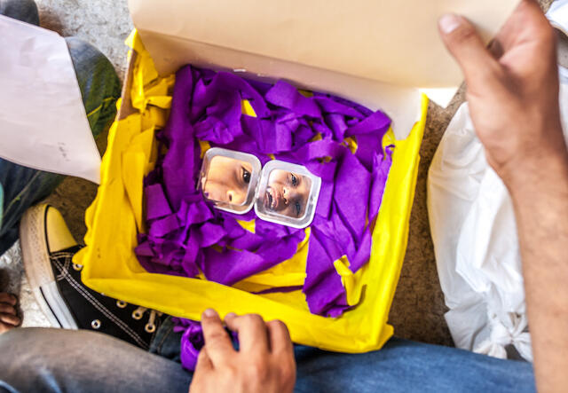 A Syrian refugee child opens a yellow box and grins when he sees a reflection of himself. 