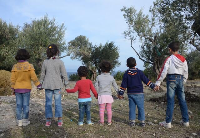 The backs of 6 refugee children stand in a line holding hands in Lesvos