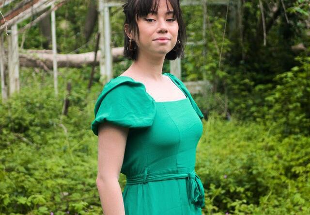 Tori Tsui looks towards the camera she is wear a green dress and is stood outside with trees in the background
