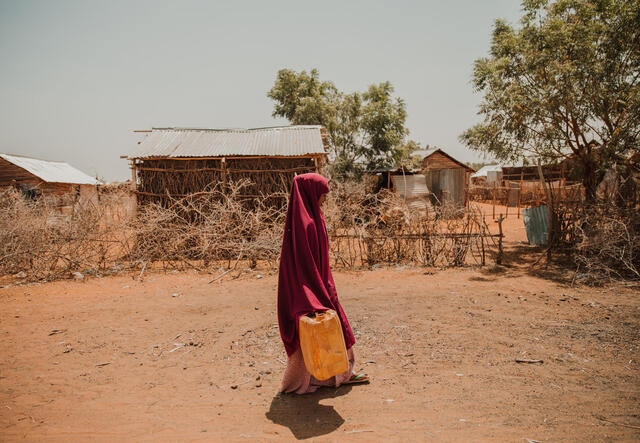 A girl in red walking on dustry road with a yellow jerry can behind her you can see houses