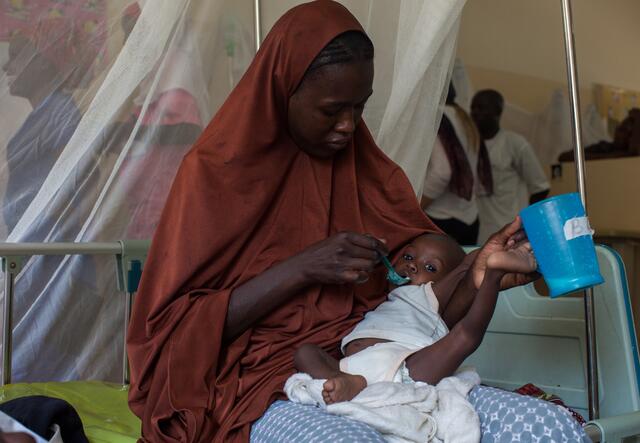 나이지리아의 IRC 진료소에서 어느 어머니가 병상에 앉아 영양 장애가 있는 아기에게 숟가락으로 치료식을 먹이고 있습니다.