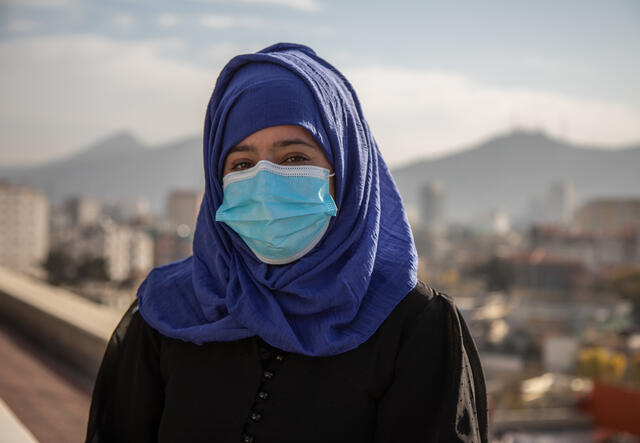 A female Afghan aid worker stands outside, with mountains in the distance. She is wearing a surgical mask to help protect against COVID-19. 