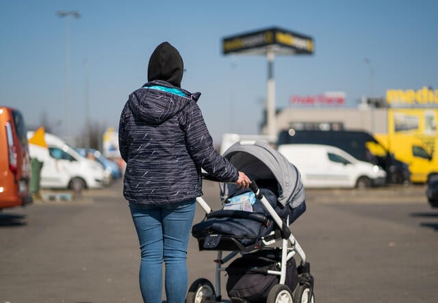 A young woman wearing a winter coat and pushing a stroller waits in a parking lot at a Polish refugee reception center.