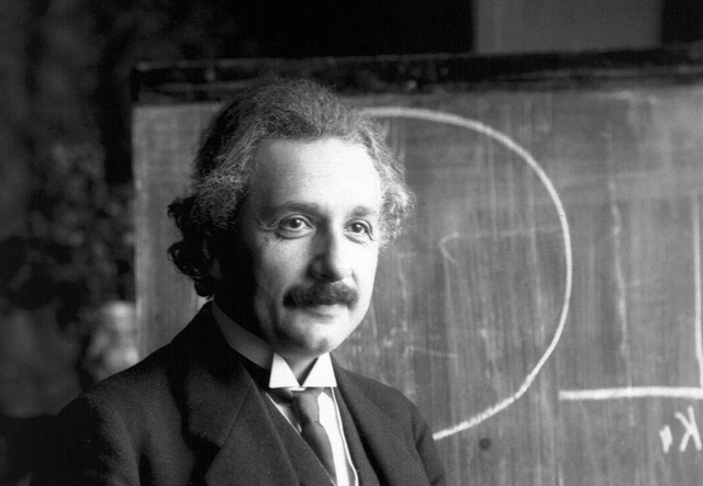 Physicist Albert Einstein stands smiling in front of a blackboard marked with chalk diagrams.
