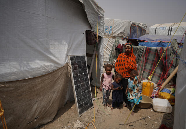 A woman and three children stand in the dirt outside of a tent, with blankets hanging behind them.