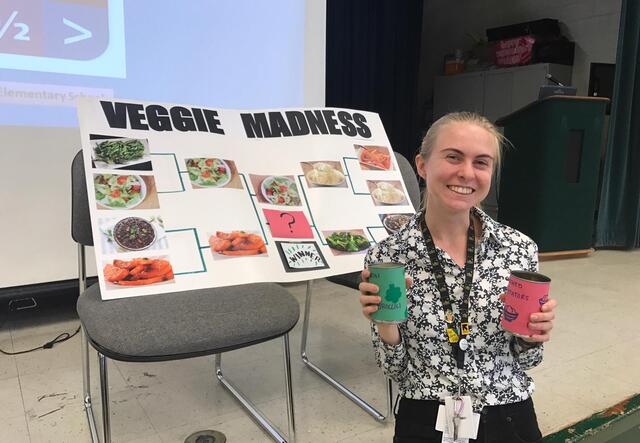FoodCorps Member Meggie stands beside her vegetable bracket poster and holds two containers for children to cast their votes.