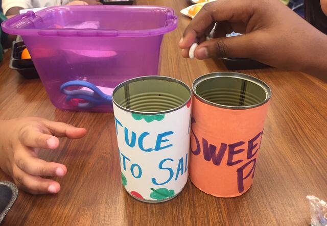Two elementary school students reach to place dried beans in containers labeled "lettuce and tomato salad" and "sweet potato."