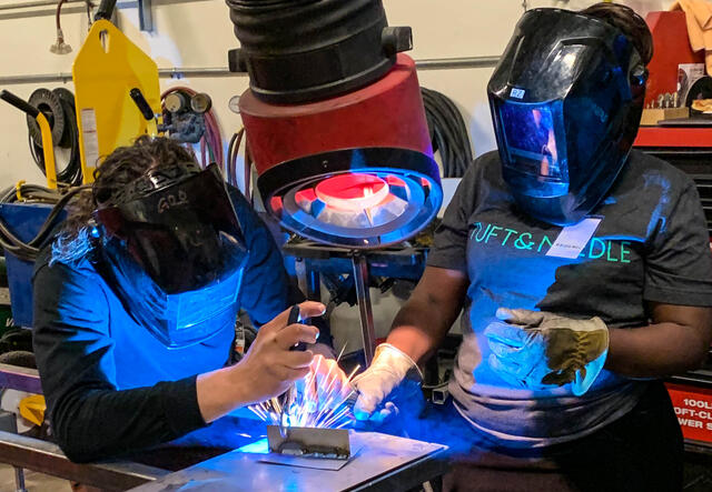 Two people wearing protective face shields are welding in a factory to gain experience with tools.