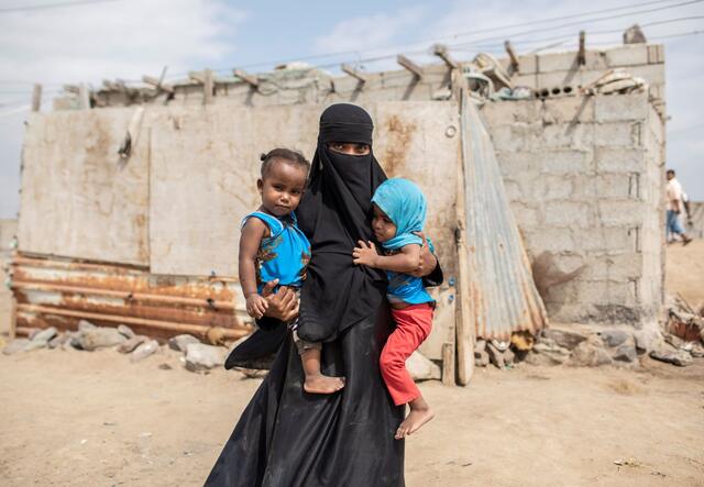 A displaced woman stands holding her toddler twin daughters outside the house they rent at a settlement for displaced people on the outskirts of Aden, Yemen.