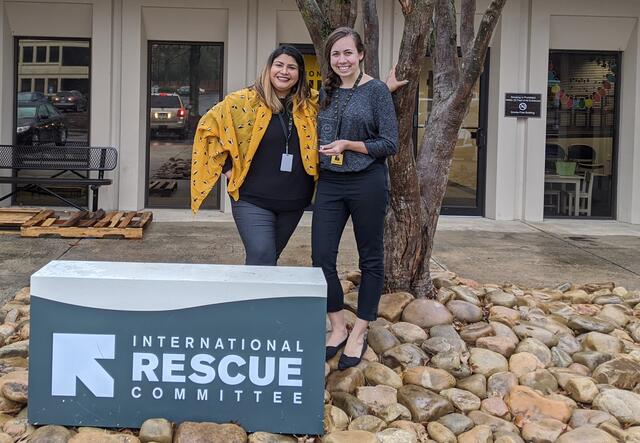 Two women stand beside a large sign that says International Rescue Committee and just in front of a medium sized tree. They are smiling and holding glass awards. They wear lanyards and work badges around their necks.