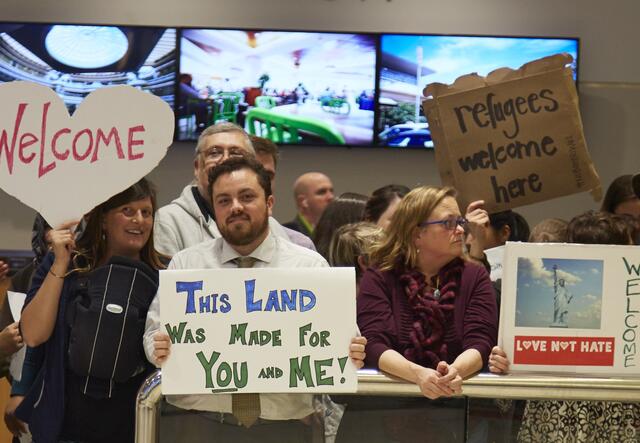 People holding different signs that say Welcome, This Land Was Made for You and Me, and Refugees Welcome stand behind an airport guard rail. 