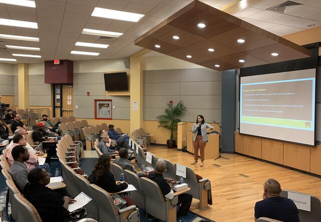 A woman stands on a stage in front of several Uber drivers, an IRC presentation is shown on the screen in the background. 