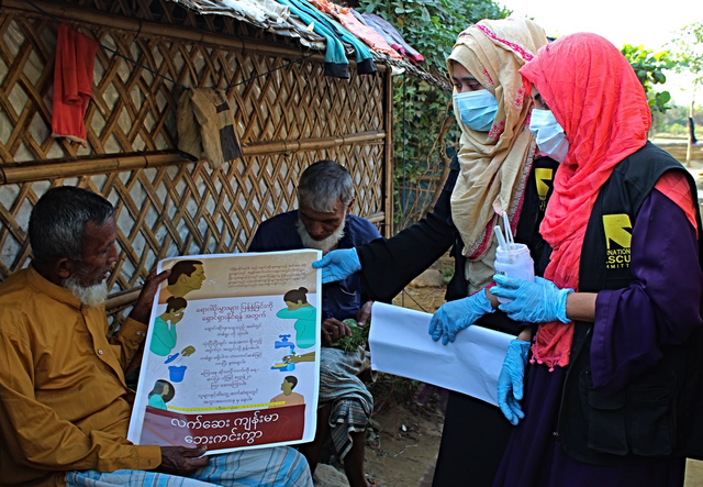 Community Health Volunteers in Bangladesh hold a poster and talk to a community member about COVID-19.