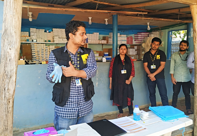 Dr. Mahmud puts on a glove to demonstrate the proper technique, while health workers and health volunteers watch him. 