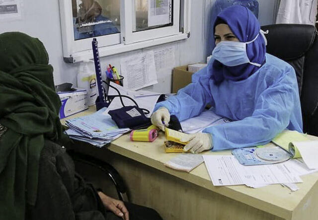 An IRC midwife wearing Personal Protective Equipment, or PPE, sits at a desk and talks with an IRC client.
