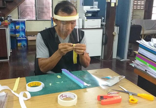 An IRC staff member in a refugee camp in Thailand sits at a desk wearing a face shield. The desk has scissors, a ruler, and tape, and he is using the supplies to create face shields.