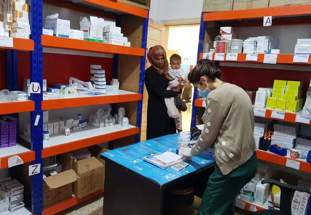 At a pharmacist in NE Syria, a pharmacist wearing a mask and gloves prepares medication while a mother holding a baby waits to get her medicine.