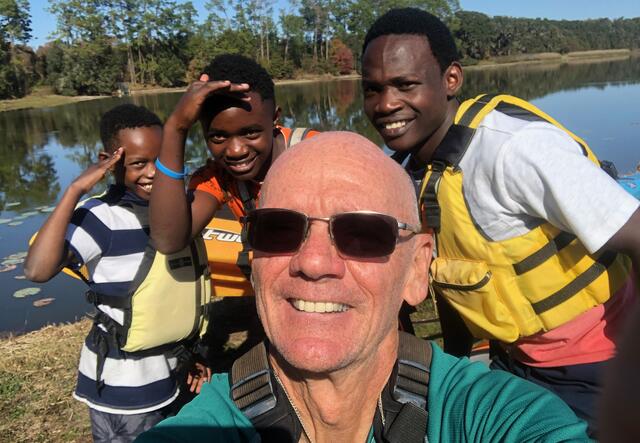 Tommy takes a selfie of him with his mentees and their brother by the river. The three young boys pose behind him and are smiling and wearing life jackets.