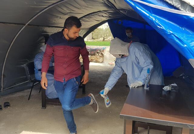 Within a tent outside of an IRC health clinic, a health worker in full PPE sprays disinfectent on the bottom of a clients shoes. Another health worker in full PPE sits at a desk behind them. 