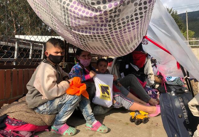 A mother with two kids sit under a makeshift tent surrounded by their luggage. They are all wearing masks and are looking directly at the camera. 