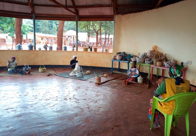 In an IRC-run safe space for women in Tanzania three woman sit on the ground six feet apart while practicing basket weazing. An IRC staff member sits on a chair facing them. 