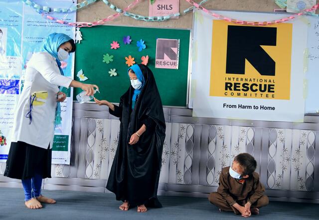 IRC hygiene promoter Sohalia Khaliqi, wearing a mask, squirts hand soap onto the hand of a child, also wearing a mask, while another child looks on. She is providing training and critical information about COVID-19 in Herat province, Afghanist