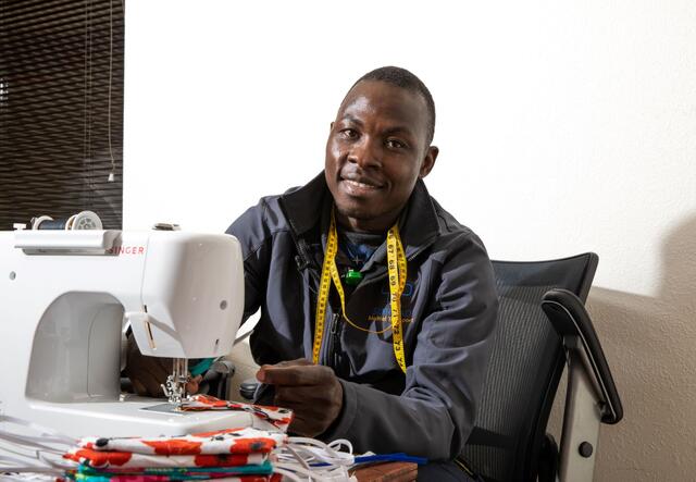 Jonathan Amissa sits at a desk with his sewing machine. He has a yellow tape measure around his neck and there is a stack of red masks on the desk. 