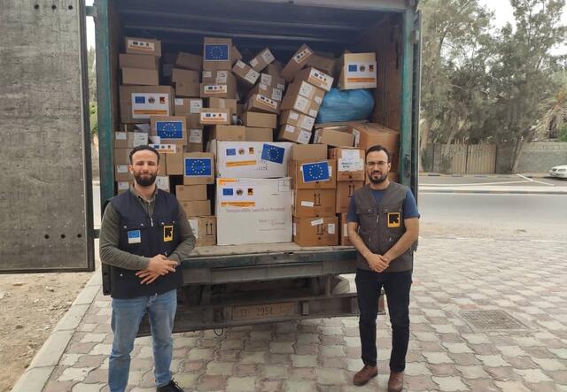Two men wearing IRC vests stand in front of a truck full of boxes of supplies to fight COVID-19, in Libya. 