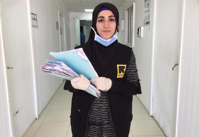 Doha Ibrahim Ammouri, holding files, walks in a hallway in an IRC reproductive health clinic. She is wearing a mask and gloves. 