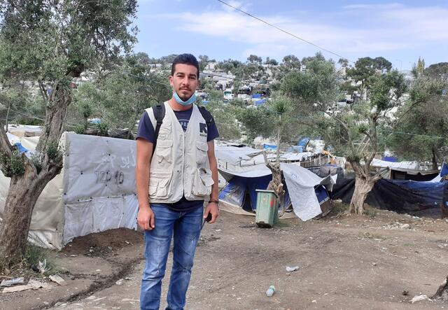 Shadi stands in front of a tree and a tent in a refugee camp. He is wearing a mask around his neck and an IRC vest.