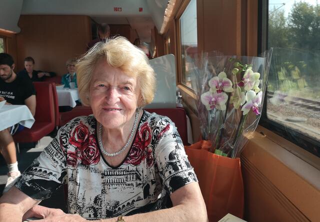 Irmgard sits at a table at a restaurant. She is wearing black and white shirt with red roses. There is a bouquet of flowers behind her. 
