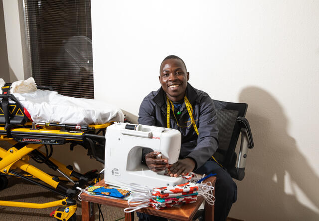 Jonathan Amissa sits at the desk where he sews masks. There is a white sewing machine and a stack of masks on the desk. 