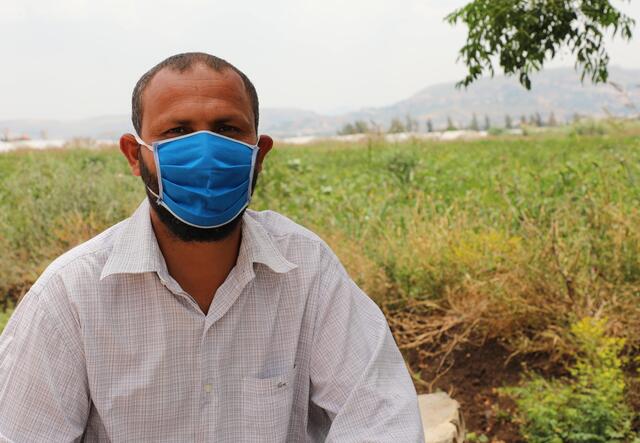 Mohamad sits on the ground and looks at the camera. He has on a white shirt and blue mask. 