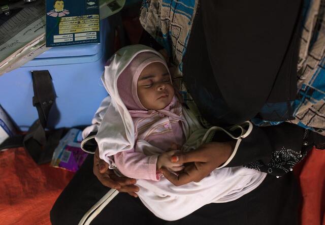 Wearing a pink onesie, baby Enkath sleeps on her mothers lap. They are inside a tent. 