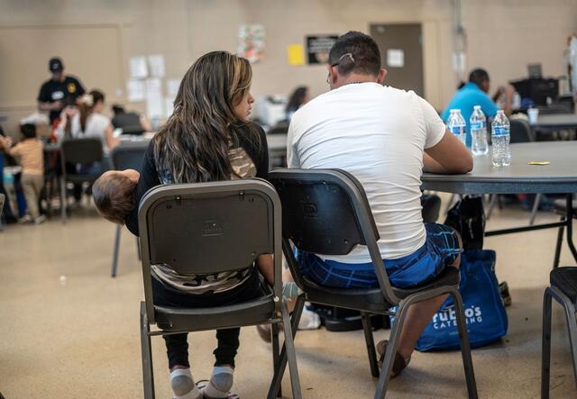 In the IRC's Welcome Center for asylum seekers, a husband and wife sit with their back to the camera as they discuss their journey as asylum seekers. The wife is holding their 2-year-old daughter. 