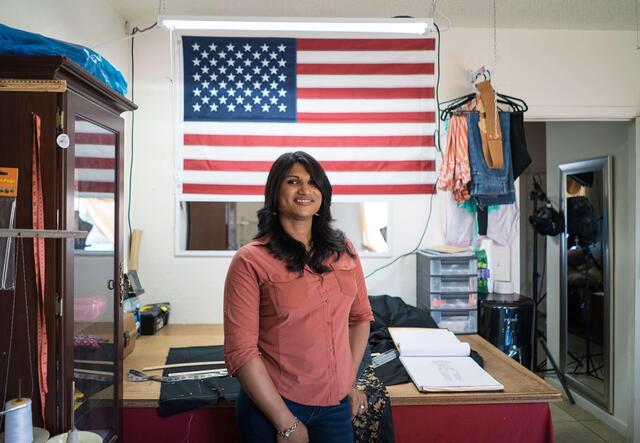 Lincy Sopall stands in her studio for her fashion design business. She is wearing a pink button down shirt and smiling. Behind her is a desk, supplies for her work, and a large American flag. 