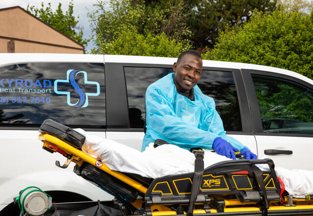 Jonathan Amissa, wearing scrubs, stands in front of a van with his hands on a stretcher. The van has a logo for Skyroad Medical Transport, his medical transportation business. 