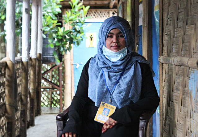 Yasmin sits on a chair on a porch in Cox's Bazar, Bangladesh. She is wearing an IRC lanyard and ID. 