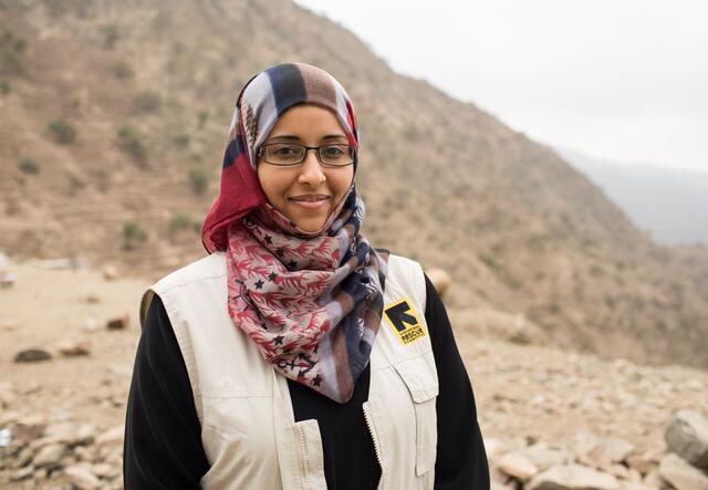 Dr. Rasha Rashed stands outside in front of a mountain in Yemen. She is wearing a colorful headscarf and an IRC vest. 