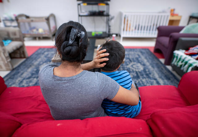 A mother sits on a red couch with her arm around her son sitting next to her. They are in a room with several chairs and a large rug. 