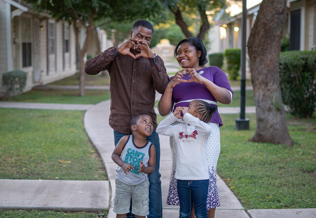 A couple and two children pose outside a row of houses and making a heart symbol with their hands.