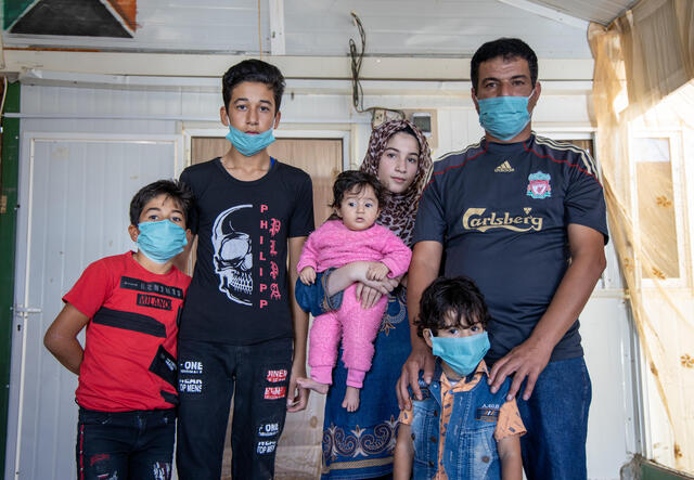 In a small tin home in a refugee camp, Manhal poses with his family: his wife holds a baby in a pink onesie, his four-year-old stands in front of him and eight-year-old and fifteen-year-old sons stands next to them. 