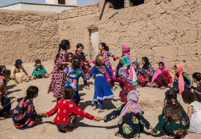 Young schoolgirls in Afghanistan hold hands sitting in a ring around a group of classmaters who are dancing.