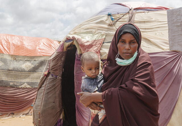 Amina stands in front of tents outside while holding their son. 
