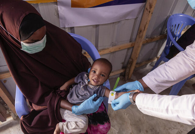 Amina holds her son while an IRC staff member wearing a white coat and blue gloves measures his arm with a color-coded armband. 