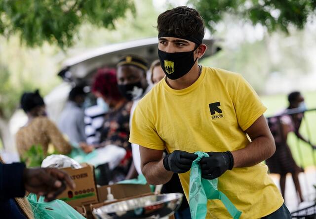 A man wearing an IRC shirt and masks, and plastic gloves is working at a farmer's market and bagging produce