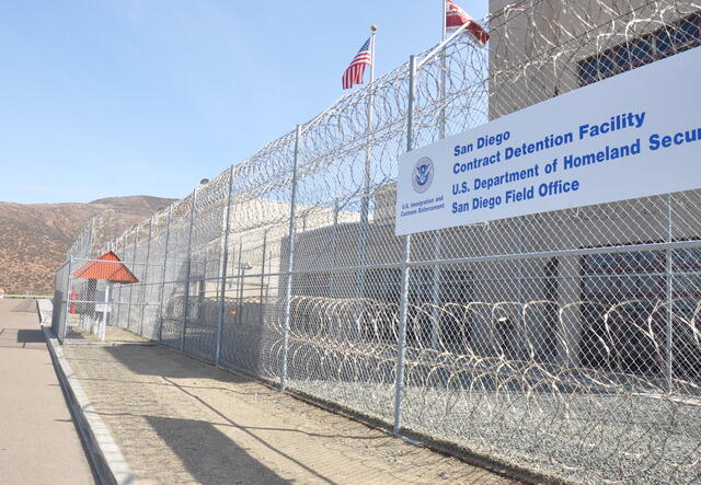 An immigrant detention center. There is a barbed wire fence and a sign that says San Diego Contract Detention Facility. 