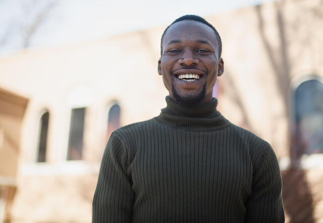 Fredrick Shema, outside and wearing a green sweater, smiles. 