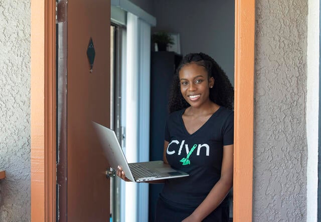 Diana Muturia stands in a doorway in her home holding her laptop and wearing a shirt with the name of her app, "Clyn" 