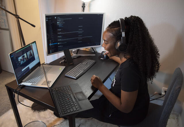 Diana sits at a desk with two laptops and a monitor. She is wearing headphones and talking to her employees at Clyn. 